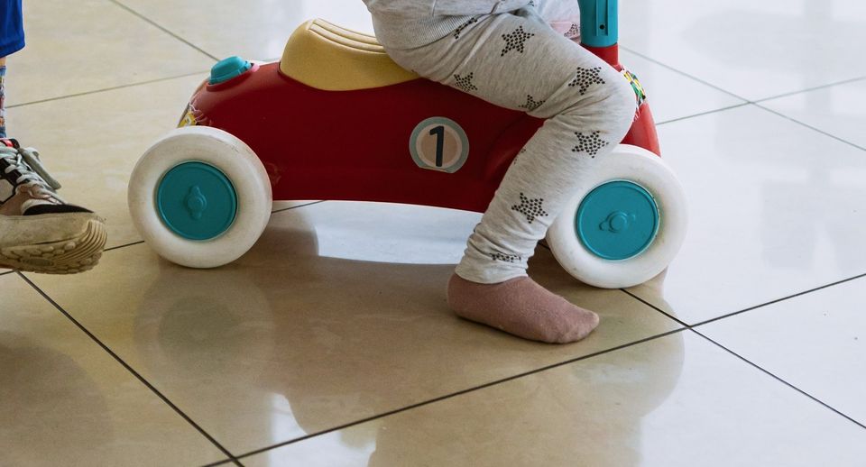 Le gambe di un bambino a cavalcioni di una macchinina rossa, un piede nudo sul pavimento